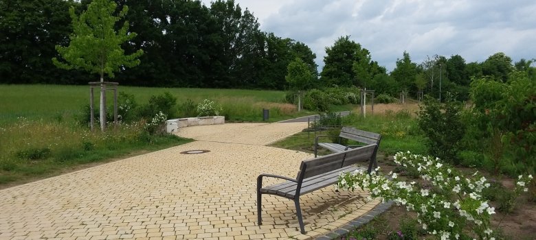 Im Vordergrund sind zwei hell geplasterte kleine Verweilbereiche mit Beton-Sitzgelegenheiten und zwei Bänken und Blumenrabatten. Im Hintergrund ist ein Feld und Bäume.