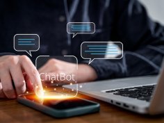 Една ръка докосва смартфон, а до него се вижда лаптоп. Виртуално се вижда балон с термина "чатбот".