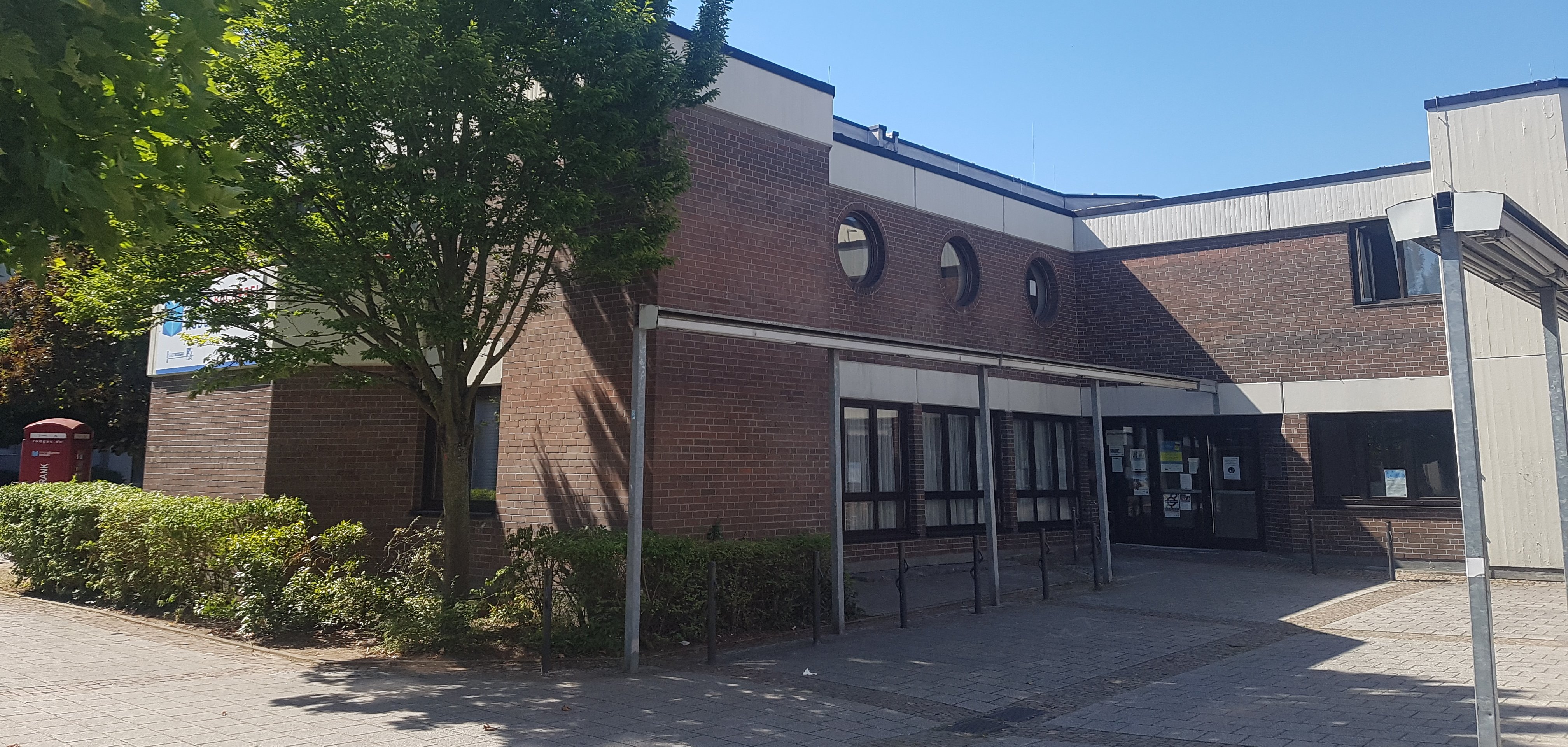 Входно пространство на социалния център Niederroden с тухлена фасада, няколко кръгли прозореца и дървета на преден план