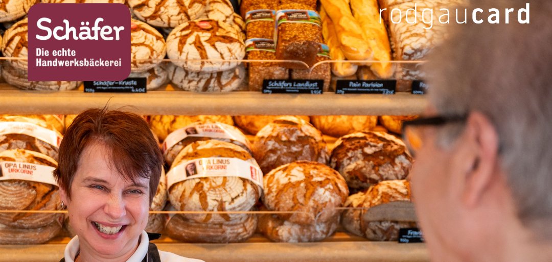 Bakery Schäfer Post - 1 Bakery with different types of bread in the background. A person in a striped shirt and apron is standing behind the counter. A customer in an orange jacket is holding a smartphone in the direction of the shop assistant. Text: 'Important information!