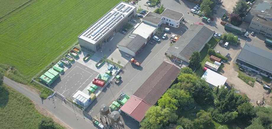 Aerial view of a recycling centre with several buildings, parking spaces and waste containers in different colours. Surrounded by meadows and trees.