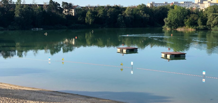 View of the lido with two floating platforms. Sandy beach and green meadow on the shore. Trees and residential buildings in the background.