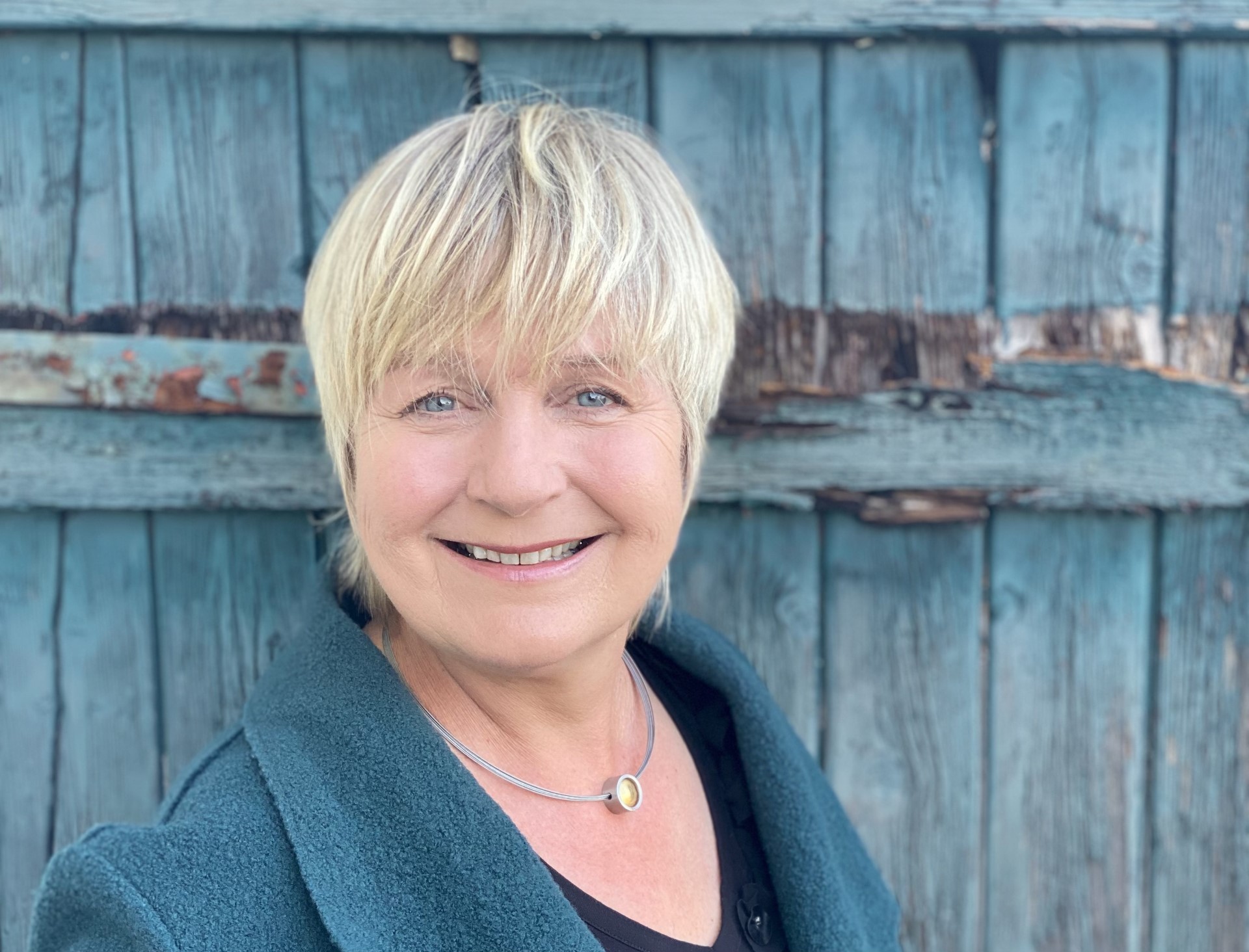 Maja Nielsen, with short blonde hair, wears a dark green coat and a necklace with a round pendant in front of a weathered blue wooden door.