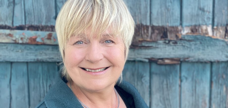 Maja Nielsen, with short blonde hair, wears a dark green coat and a necklace with a round pendant in front of a weathered blue wooden door.