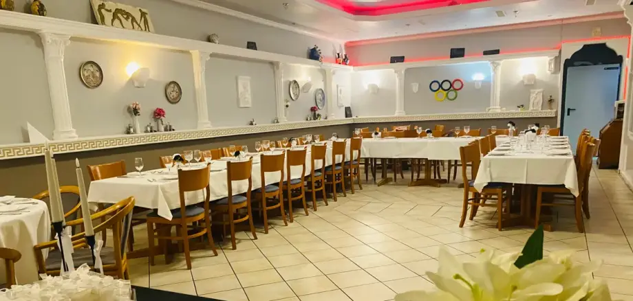 Olympos - partner of the rodgaucard Large, festively decorated dining room with several long tables, wooden floor and white tablecloths, decorated with flowers and wall clocks.
