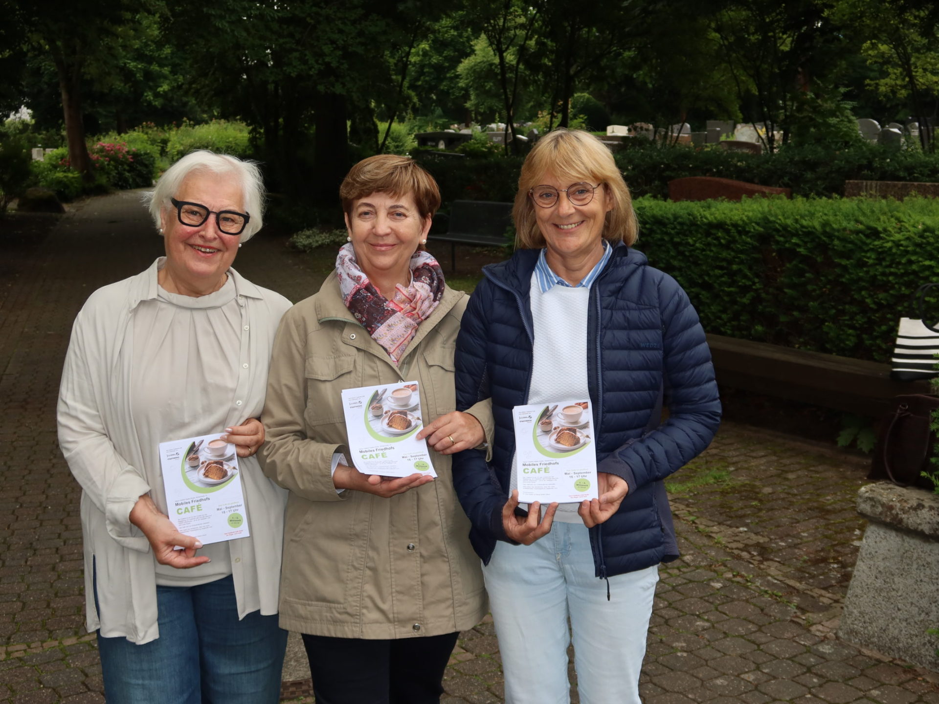 Three women are standing on a paved path in a park, each holding an information leaflet with the title 'Café'.