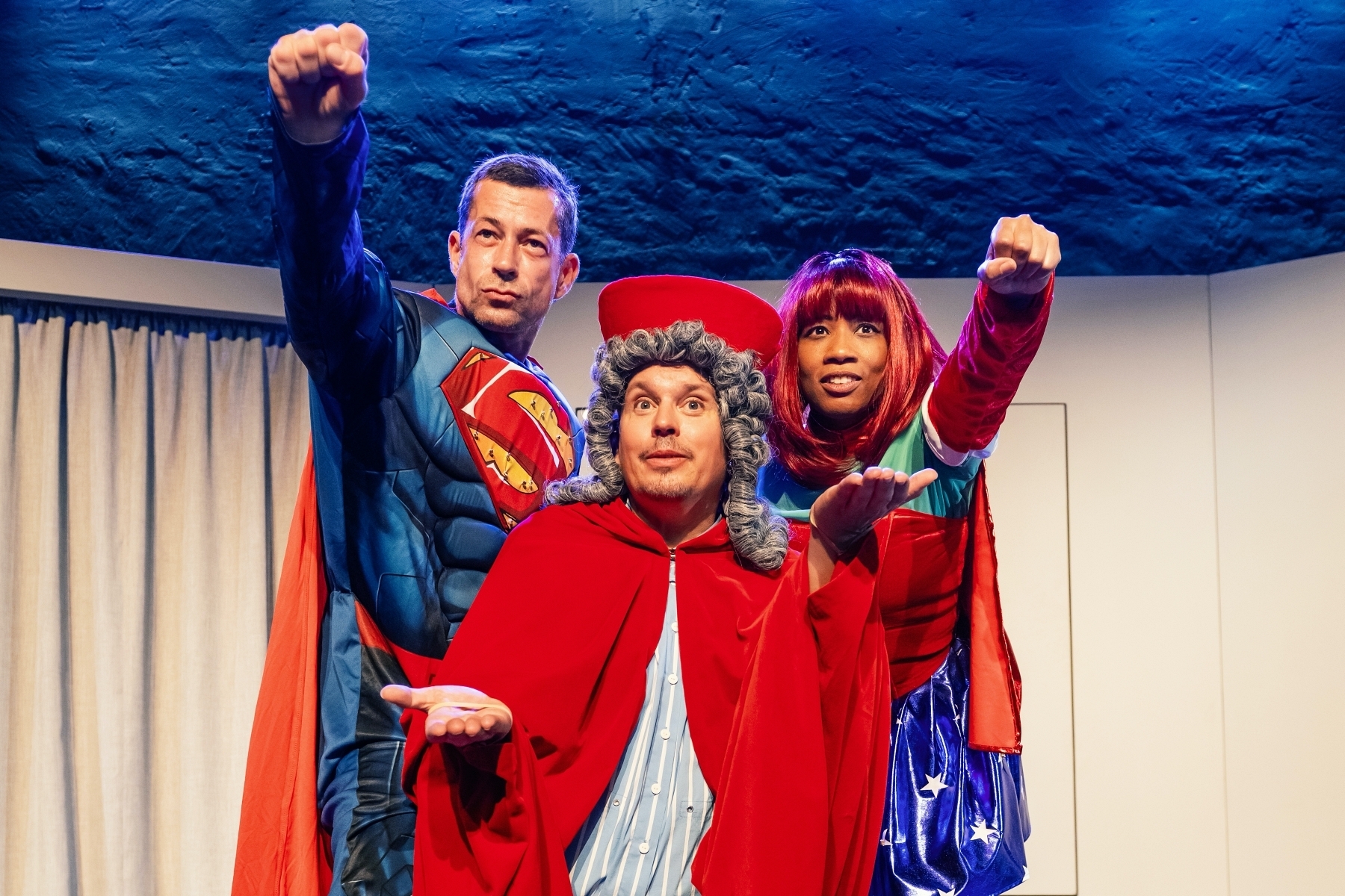 The SchMERZtherapie Three people on a stage: one in a red cape and grey wig, two in superhero costumes with red capes, each holding a fist out in front of them.