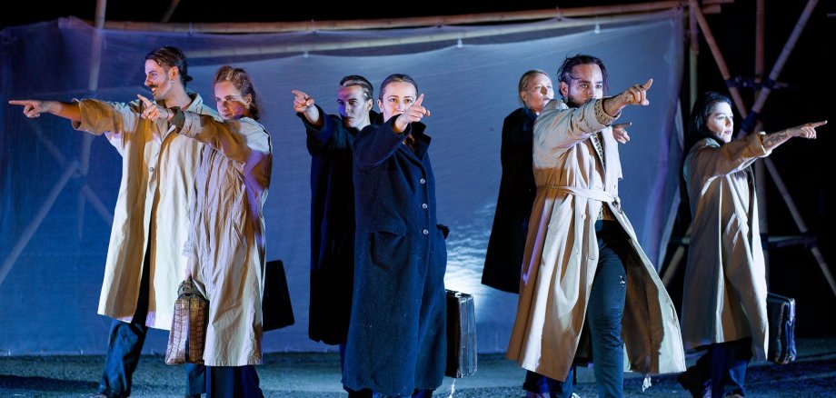 The stage is dark blue. Six people in long, light-coloured coats stand in a row and point their fingers in different directions. Some are holding bags in their hands.