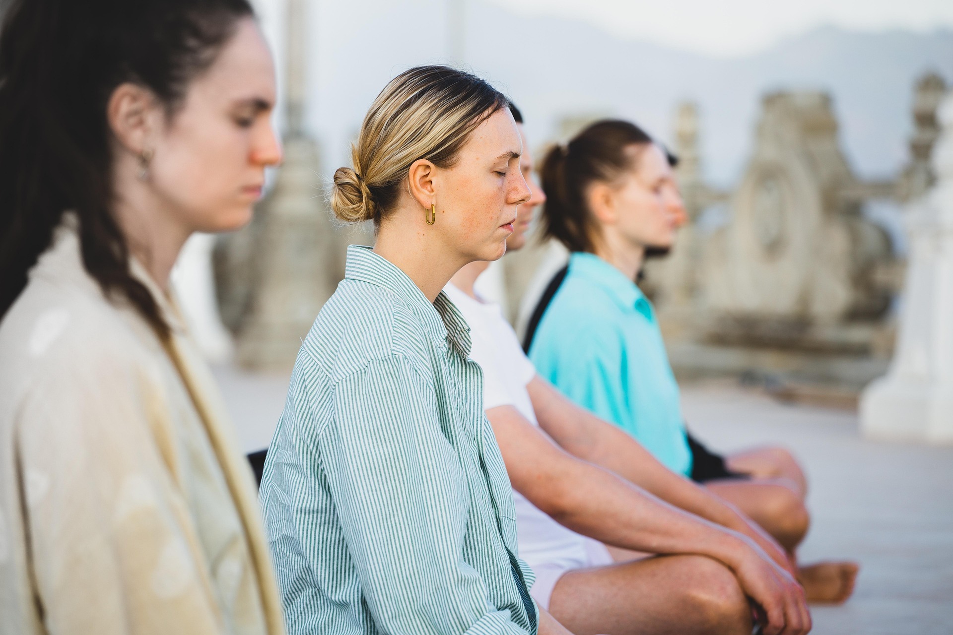 Meditation Four people sit in a row next to each other, all in profile, in front of a stone wall with decorative elements, outside and meditate.