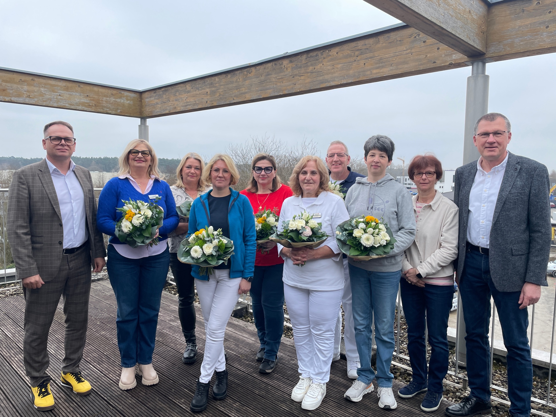 Group of eleven people standing on a wooden terrace in front of a landscape, several women holding bouquets of flowers, two men standing at the sides.