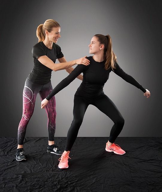 Two women in sportswear; one is in a deep squat with her arms outstretched, whilst the other has her hand on her shoulder to support her