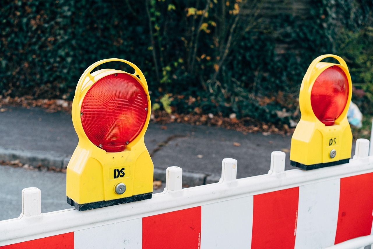 Traffic obstruction Two yellow warning lights with red light on a white barrier with red and white stripes. Green plants are visible in the background.