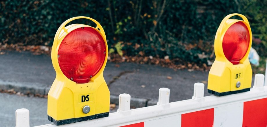 Two yellow warning lights with red light on a white barrier with red and white stripes. Green plants are visible in the background.