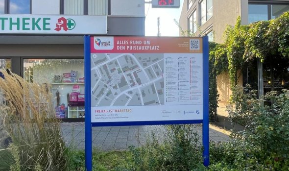 Information sign with city map and text 'ALL ABOUT PUISEAUXPLATZ' in front of a building with a pharmacy and pharmacy sign