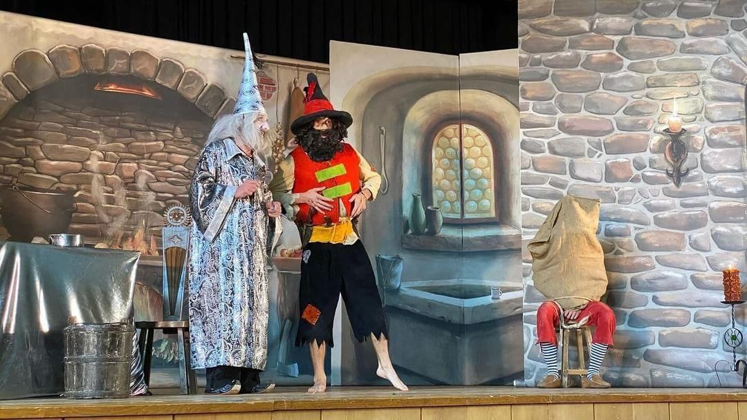Theatre scene with three people in costumes: a magician with a long white beard and pointed hat, a figure with a red shirt, black hat and beard, and a seated person with a sack over his head in red trousers