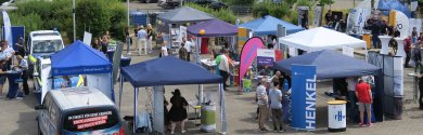 Outdoor area of an education fair with several stands, visitors and a Henkel stand under a blue sky