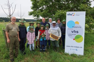 Group of children and adults outdoors, one person wearing beekeeper's clothing and holding a honeycomb, next to a banner of the Rodgau adult education centre