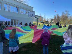 Children and carers hold a large round cloth with balls in their hands and twirl the balls in the air. In the background is the Dudenhofen youth centre with its outdoor area and basketball hoop.
