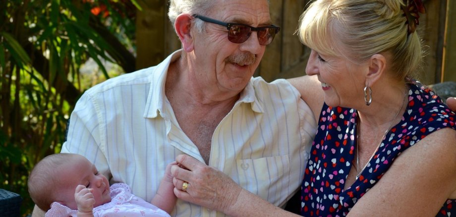 Elderly couple sitting outside, the man holds a baby in pink clothes in his arms, the woman touches his chest and looks at him