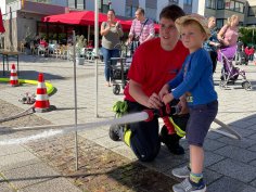 Child with straw hat holding fire hose, supported by fireman in red T-shirt, at outdoor children's party