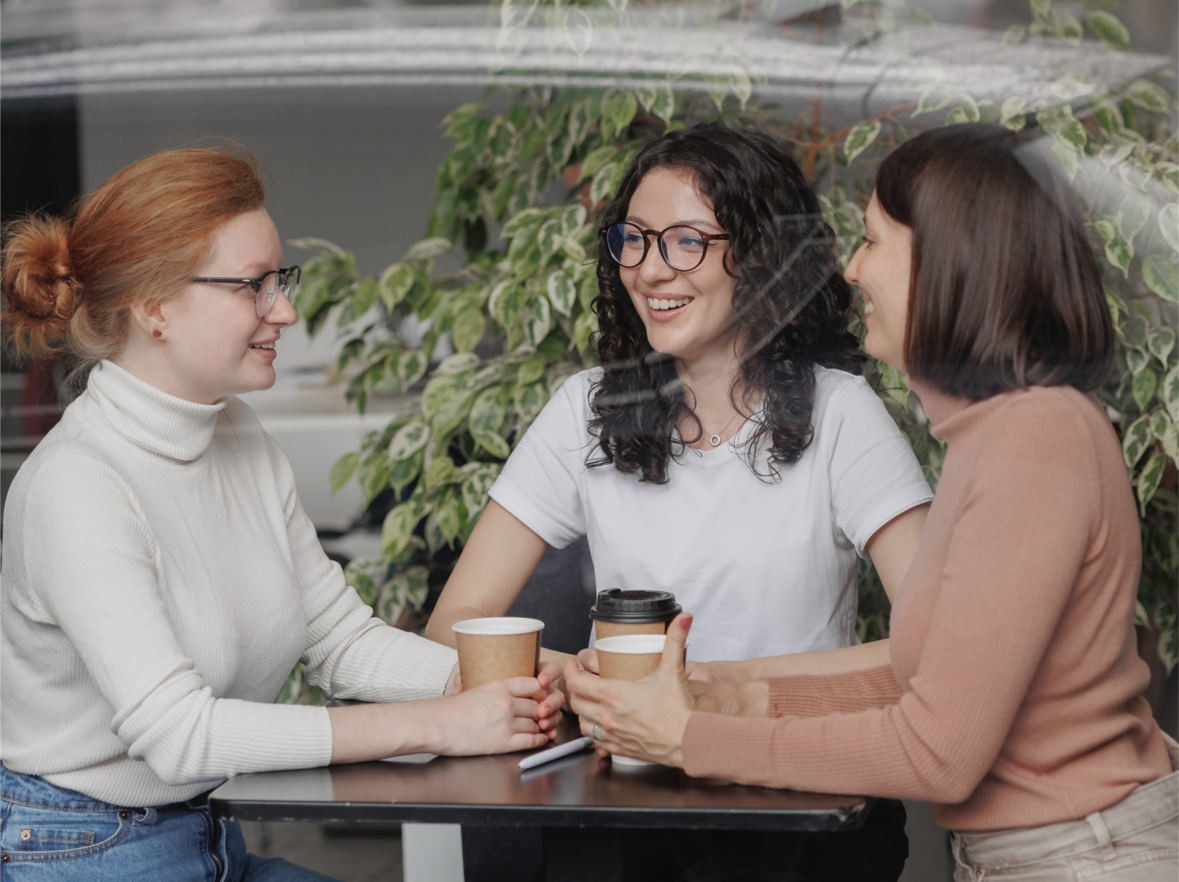 Mum Connection Three women sitting at a table, holding paper cups and chatting, a green plant in the background