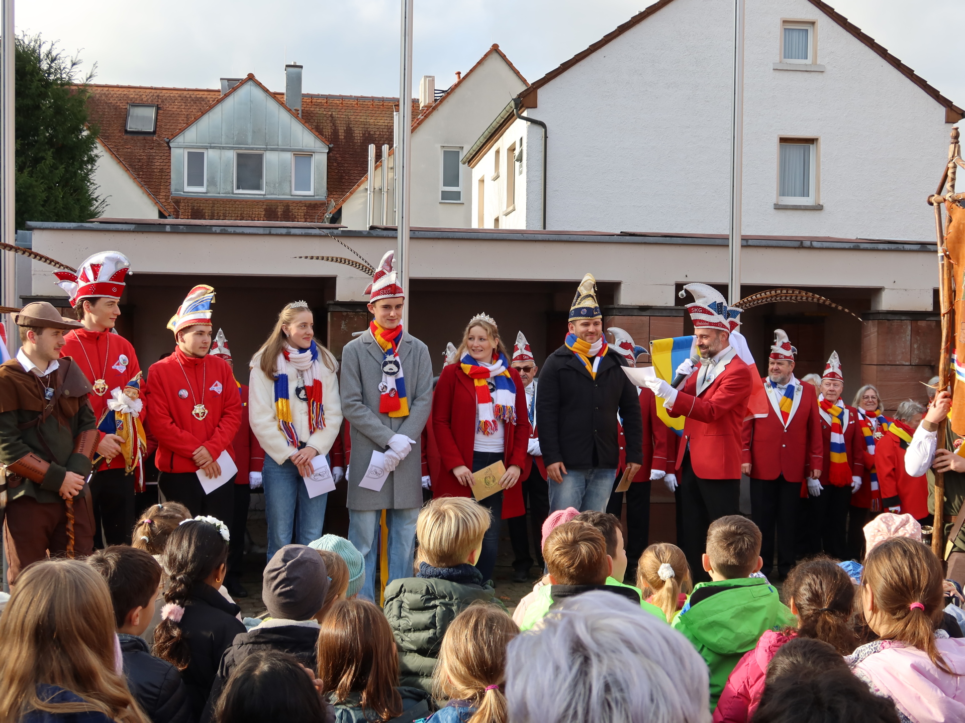 People in colourful carnival costumes and sashes stand in front of an audience on the town hall square with houses in the background.