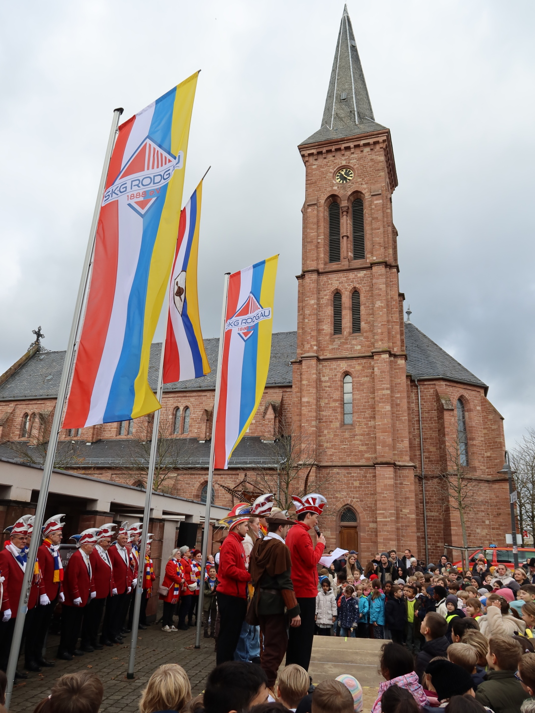 The carnival flags are hoisted, the officials stand on the stage, surrounded by the audience in the background is the church of St Nicholas.