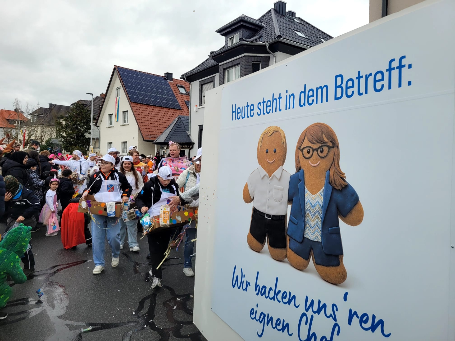 City employees with the Mayor and First City Councillor in costume at the carnival parade behind a large sign with the Mayor and First City Councillor as two gingerbread figures and the text 'Today the subject says: We're baking our own boss'.