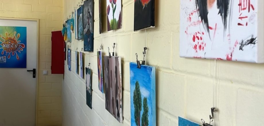 Works of art from JugendArt hang on a white wall in a corridor, including a portrait of a woman with black hair and a red background