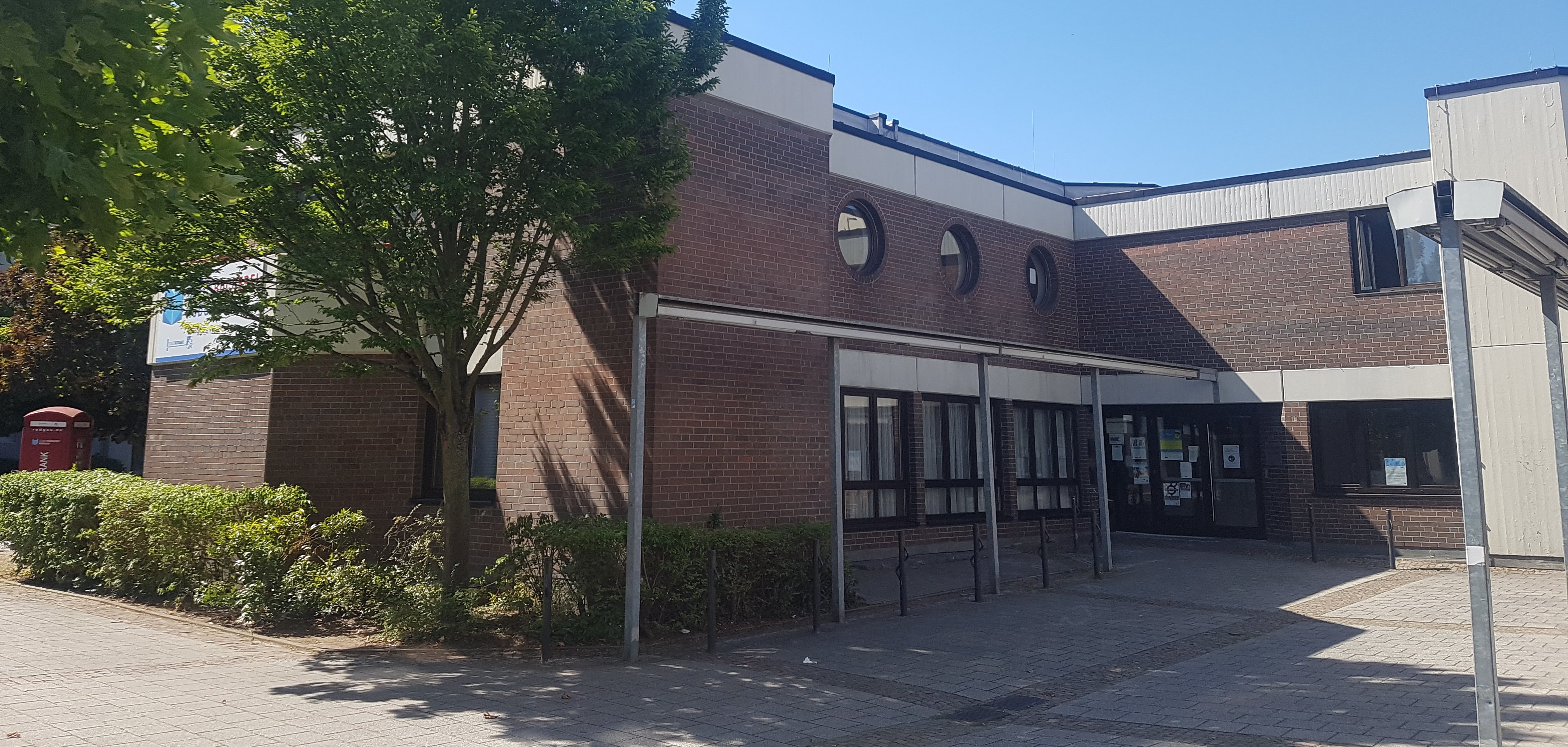 Entrance area of the Niederroden social centre with brick façade, several round windows and trees in the foreground
