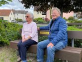 A lady and a gentleman are sitting on a bench in a square in Hainhausen with lots of greenery.