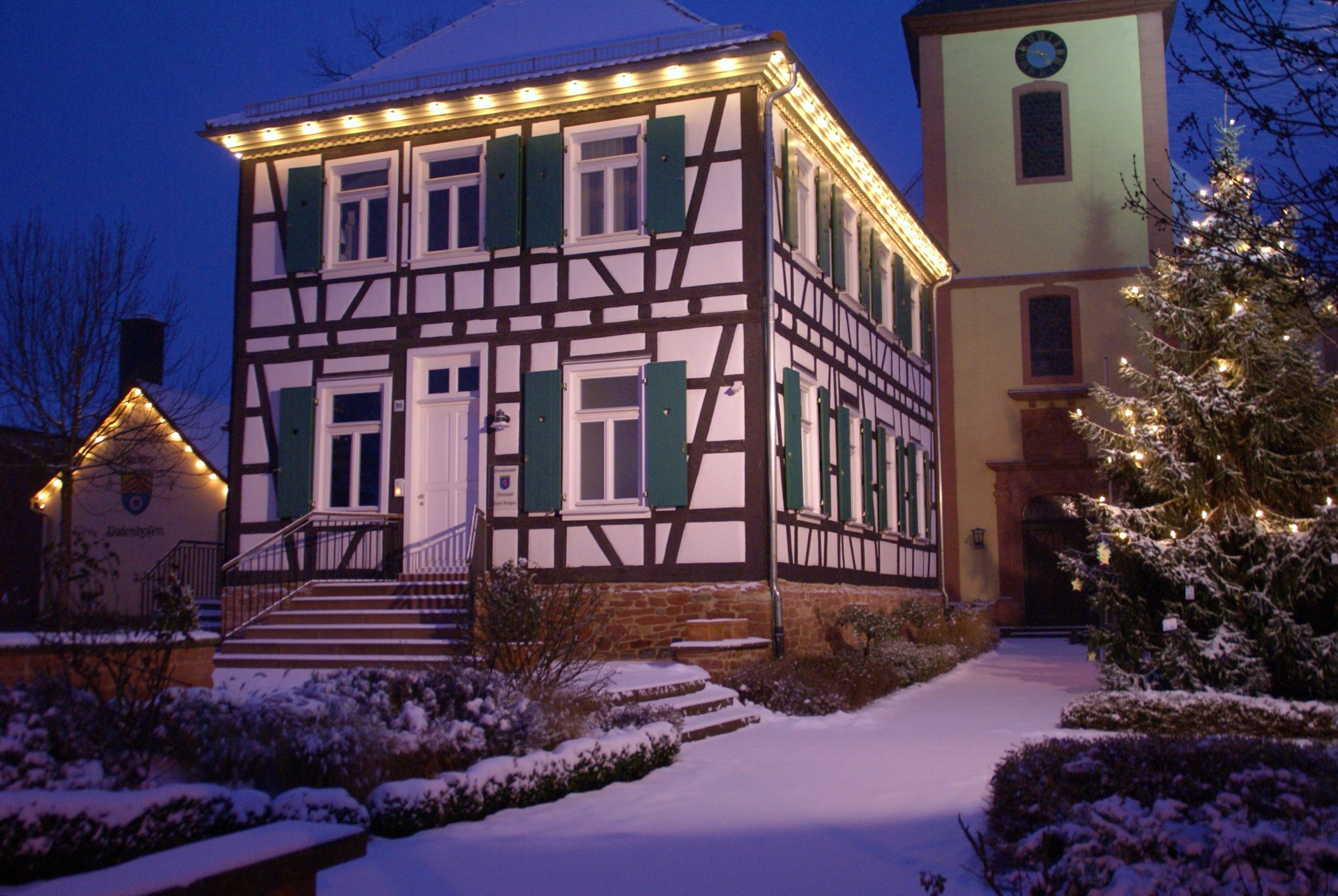 Rector Geissler House with green shutters and Christmas lights, snow-covered garden and illuminated Christmas tree in front, church tower in the background at dusk.