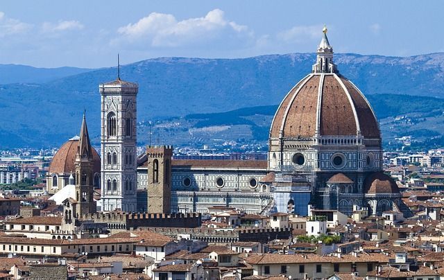 Vista de la cúpula y el campanario de la catedral de Florencia con un telón de fondo de montañas y una ciudad con tejados de tejas rojas