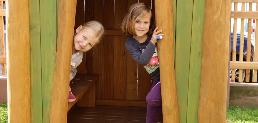 Casa de juegos de madera con paredes pintadas de verde y una puerta abierta. Dos niños están de pie en la entrada, uno mira hacia fuera. Una pequeña escalera conduce a la puerta.