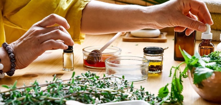 Diversas hierbas silvestres y hierbas secas Mano de una persona con camisa amarilla que sostiene un pequeño frasco de cristal con cuentagotas sobre una mesa de madera con varias hierbas silvestres, pequeños tarros de líquidos y un recipiente blanco con hierbas secas.