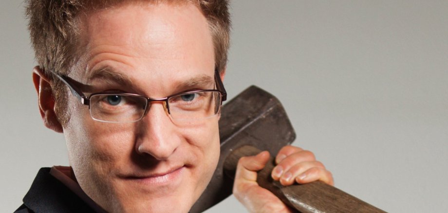 Persona con el pelo corto y rubio que viste un traje negro y una camisa rosa claro y sostiene un hacha pesada sobre el hombro