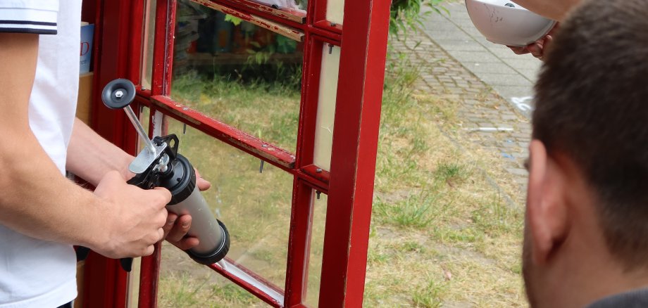 Acción 16 Plus Un joven sujeta una puerta de madera roja mientras otra persona con un brazo tatuado estabiliza la puerta y una tercera sujeta un smartphone en un trípode
