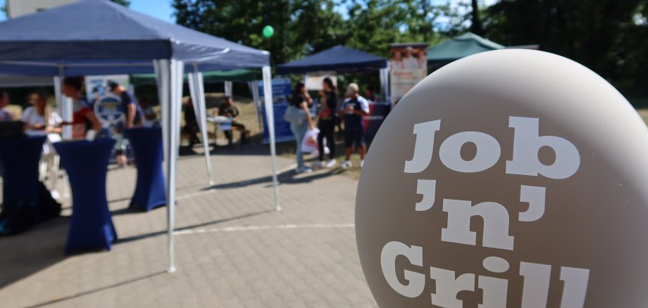 Globo con la etiqueta "Job 'n' Grill" ante los stands y visitantes de una feria de educación al aire libre