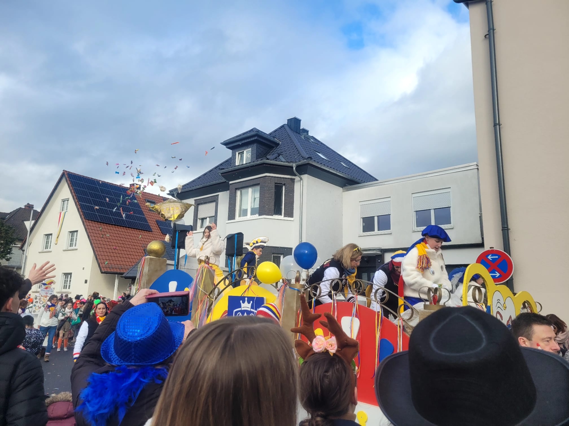 Princesa y príncipe infantiles con disfraces festivos en una colorida carroza en el desfile de carnaval frente a las casas, rodeados de espectadores que les aclaman con sombreros y globos.