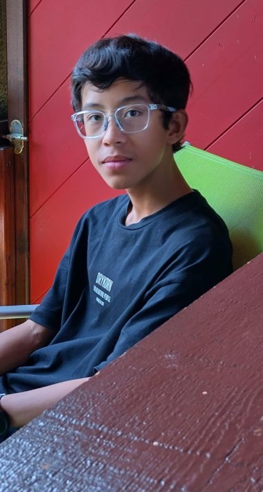 Un joven con el pelo corto y oscuro y gafas transparentes está sentado en una silla verde frente a una pared de madera roja