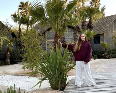 Una mujer, vestida con unos pantalones blancos holgados y un jersey rojo oscuro, se encuentra de pie sobre una pasarela de madera y sostiene una palmera en un jardín tropical con casitas de madera al fondo.
