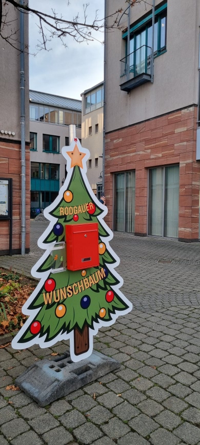 Una colorida placa en forma de árbol de Navidad con un buzón rojo, con la inscripción "RODGAUER WUNSCHBAUM", se alza en una plaza pavimentada frente al edificio del ayuntamiento.