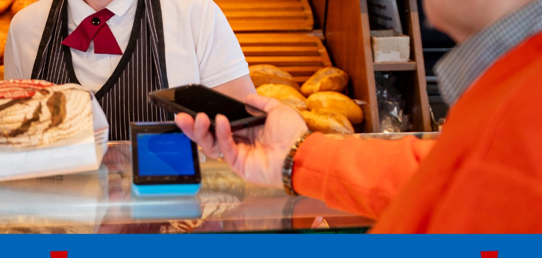 Boulangerie avec différentes sortes de pain en arrière-plan. Une personne en chemise rayée et tablier se tient derrière le comptoir. Un client vêtu d'une veste orange tient un smartphone en direction de la vendeuse. Texte : 'Information importante !