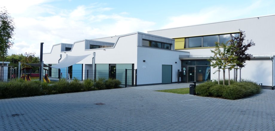 Bâtiment blanc moderne du centre familial avec une esplanade pavée, plusieurs fenêtres et un petit arbre devant.
