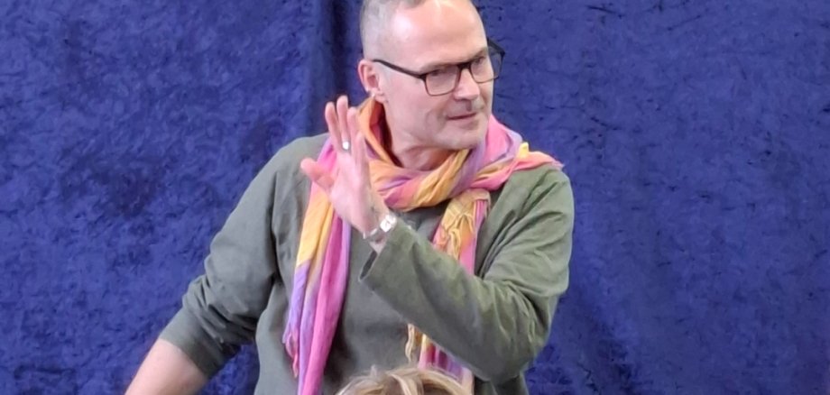 Le conteur Christian Mayer-Glauninger est assis par terre devant une couverture bleue, porte une écharpe colorée et gesticule de la main droite devant un groupe d'enfants.