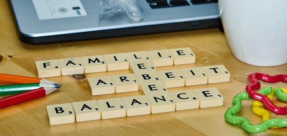 Des pièces de Scrabble forment les mots 'FAMILLE', 'TRAVAIL' et 'BALANCE' sur une table en bois à côté d'un ordinateur portable, d'une tétine, de crayons de couleur, d'un gobelet de café et de jouets colorés.