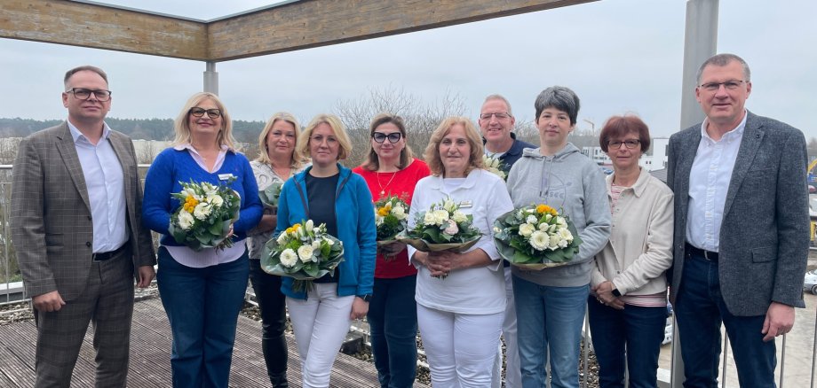 Groupe de onze personnes debout sur une terrasse en bois devant un paysage, plusieurs femmes tenant des bouquets de fleurs, deux hommes debout sur les côtés.