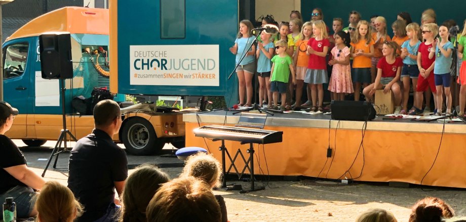Chorale d'enfants chantant sur une scène devant un camion bleu portant l'inscription 'DEUTSCHE CHORJUGEND #chantez ensemble nous sommes plus forts'.