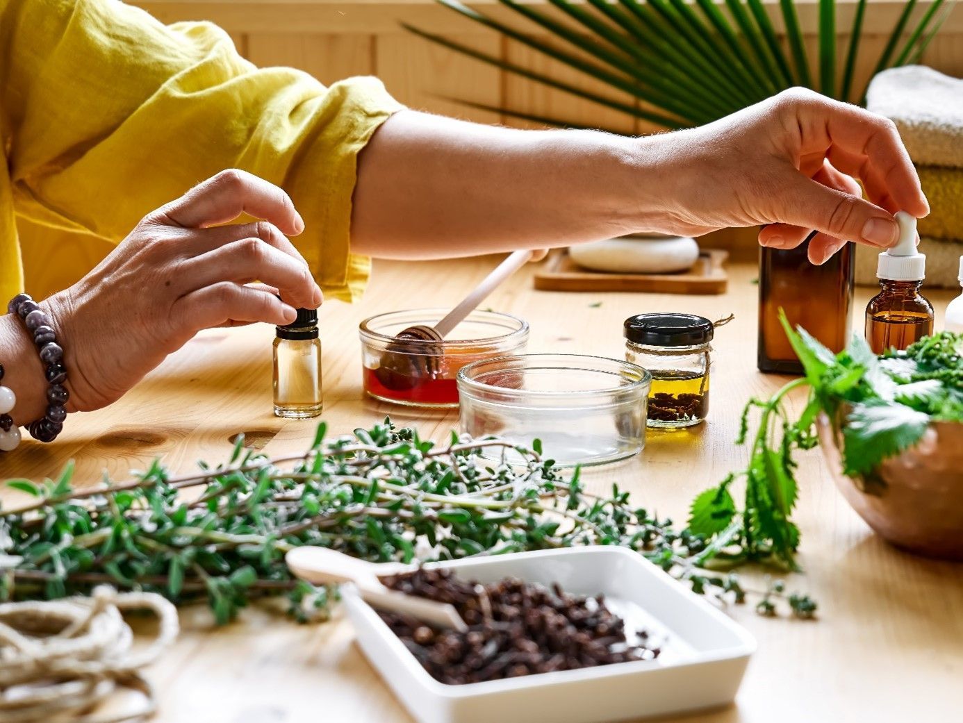 Main d'une personne en chemise jaune tenant une petite bouteille en verre avec compte-gouttes au-dessus d'une table en bois contenant diverses herbes sauvages, des petits verres remplis de liquide et un récipient blanc contenant des herbes séchées.