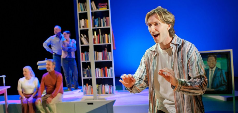 Pièce de théâtre allemande Un homme vêtu d'une chemise rayée gesticule avec animation sur scène, devant un groupe de personnes assises et debout, ainsi qu'une bibliothèque et un téléviseur sur lequel apparaît l'image d'un homme